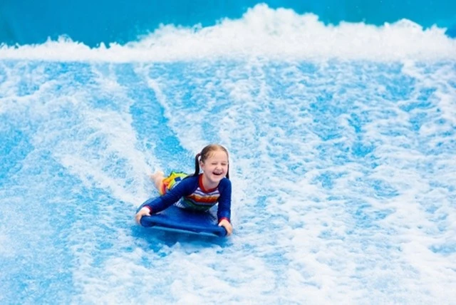 Little girl surfing in beach wave simulator attraction in water amusement park of tropical resort.